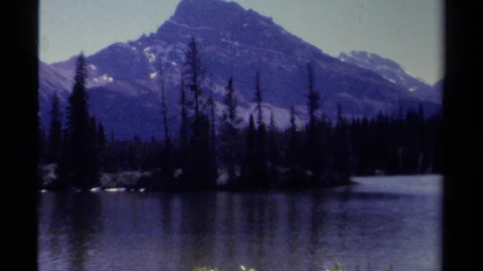 British Columbia mountain scenery on 8mm film, with dark evergreen trees, open water, and a large peak rising behind the forest.