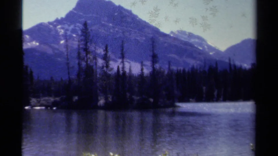 Soft 1979 8mm view of a forested British Columbia shoreline, calm water, and layered mountain peaks in the distance.