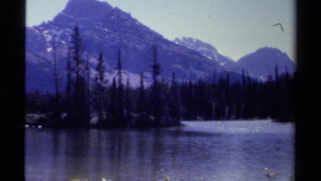 1979 British Columbia 8mm frame showing a calm alpine lake, dark evergreen shoreline, and snow-dusted mountain peaks.