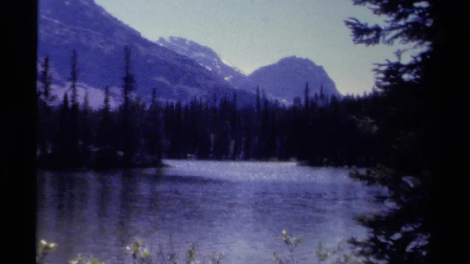 1979 British Columbia lake view with mountains behind a dark evergreen shore, recorded in soft blue-shifted 8mm color.