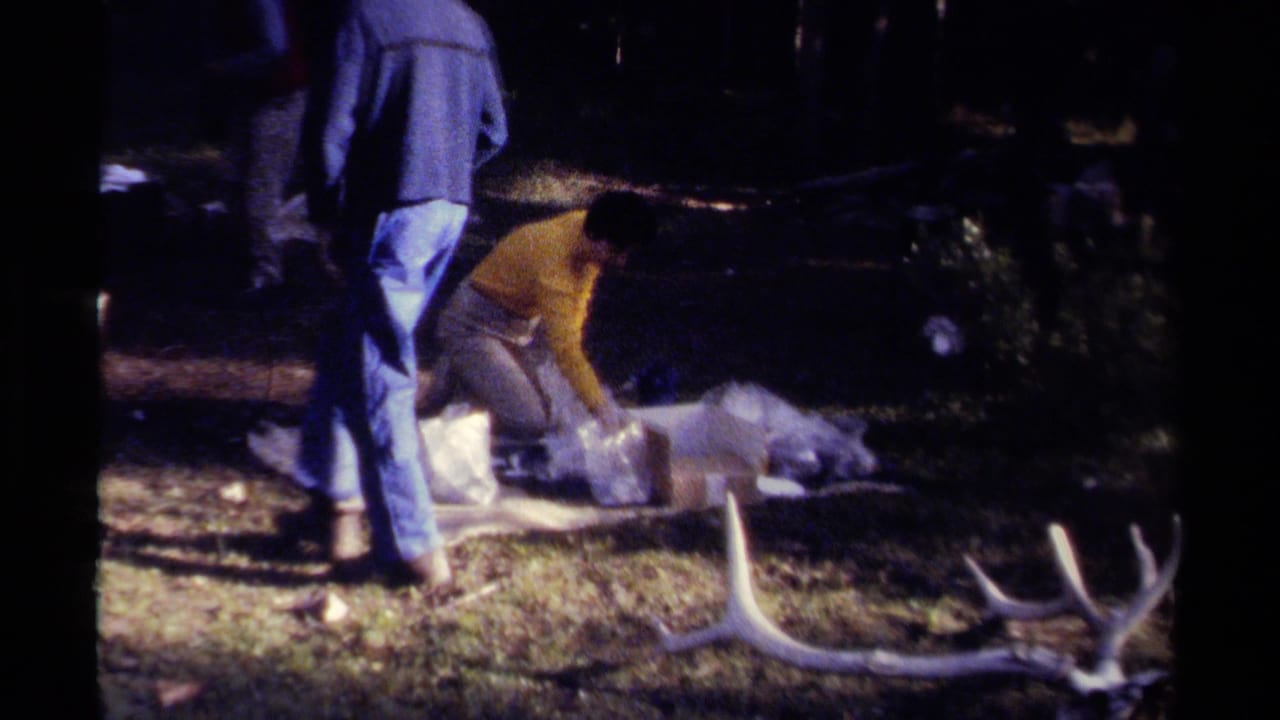1979 British Columbia 8mm frame showing adults in casual clothing around a deer carcass laid on a tarp in a grassy shaded area.