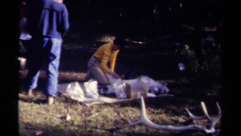 1979 British Columbia home-movie frame with a kneeling person in yellow beside a deer carcass, while another adult stands nearby.