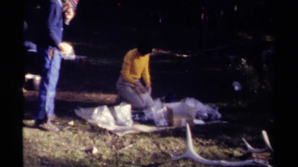 1979 British Columbia 8mm frame showing adults continuing outdoor deer processing, with antlers lying across the lower foreground.