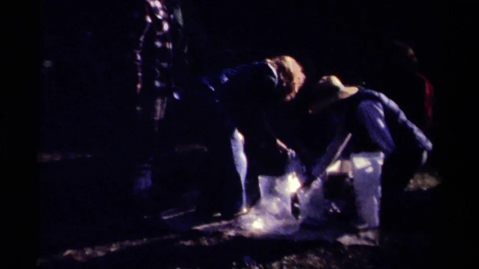 A darker 1979 British Columbia 8mm frame shows adults bending over the tarp and carcass during an outdoor deer-processing moment.