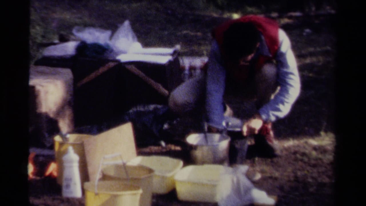 In 1979 British Columbia home-movie footage, a person in a red vest crouches over metal pots and yellow containers at a forest campsite.