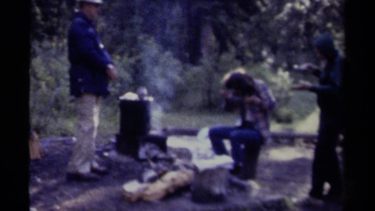 Three people around a smoky campfire and cooking pot in a wooded campsite.