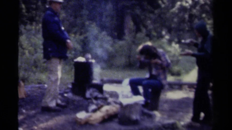 Three adults appear near a smoking campfire and black cooking pot in a wooded British Columbia campsite, consistent with the 1979 metadata.