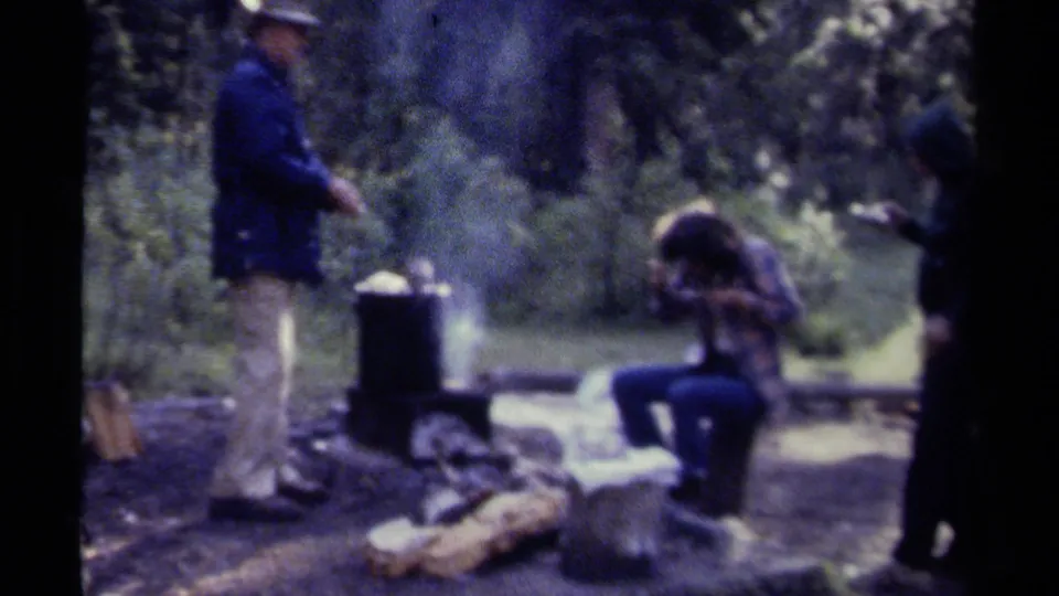 A man stands by a smoking camp pot while another person sits nearby, with a third figure partly visible at the edge of the forest campsite.