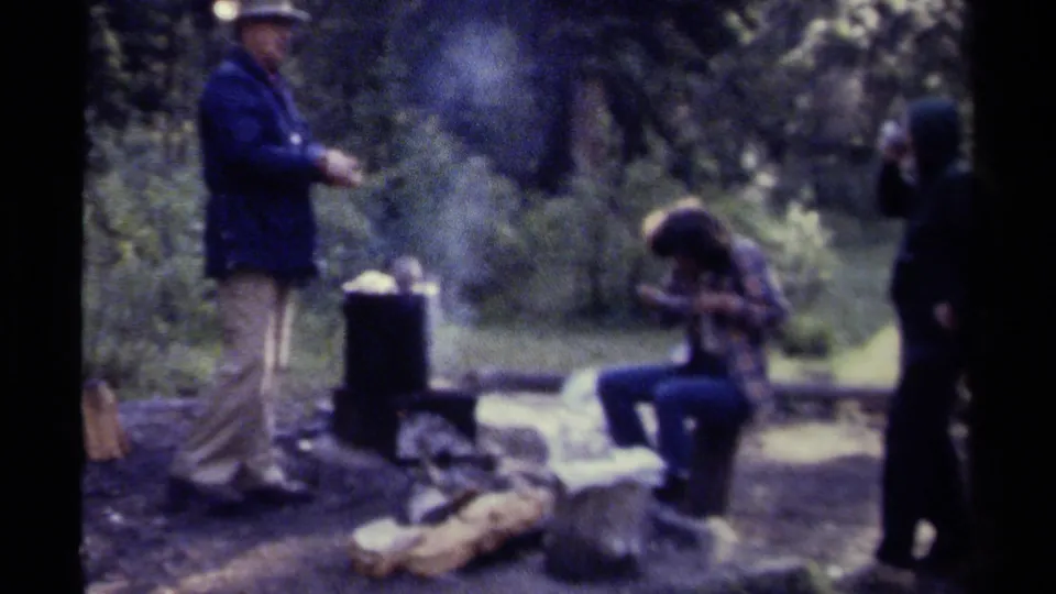 Campers remain near a smoky outdoor cooking pot and logs in a British Columbia forest setting, captured with soft 8mm home-movie texture.