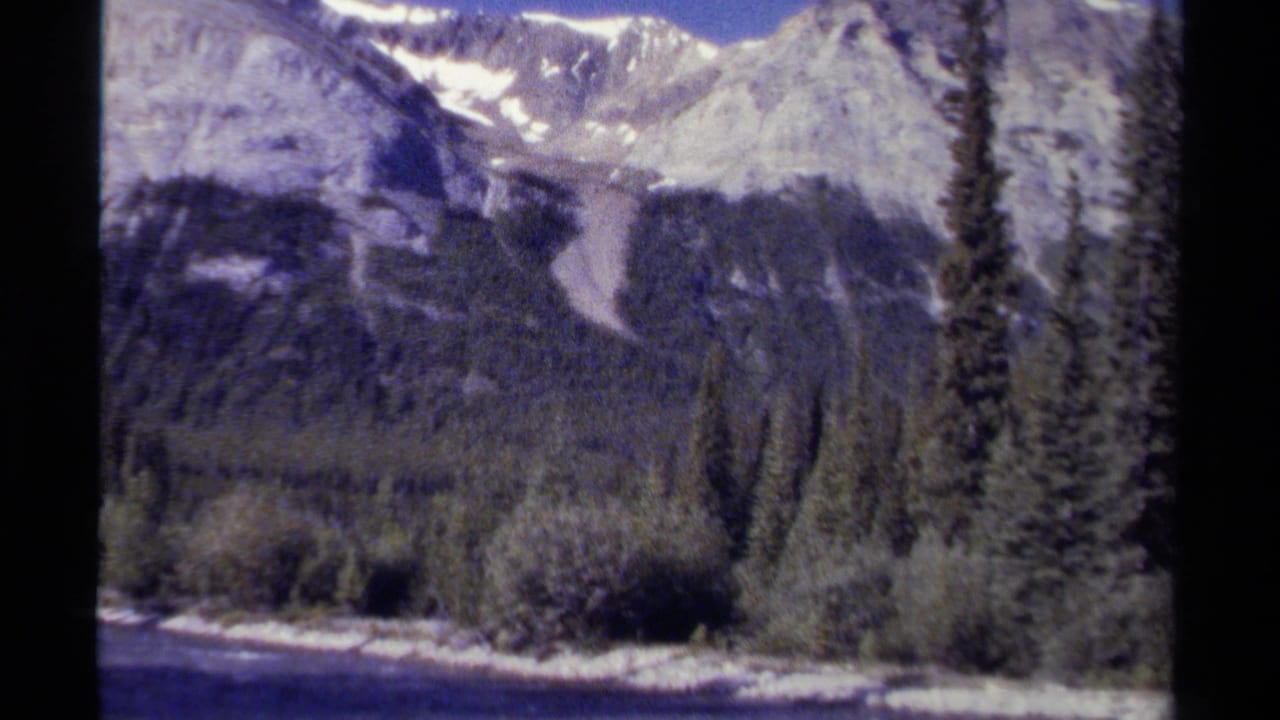 Snow-dusted mountains above evergreen forest and a riverbank.