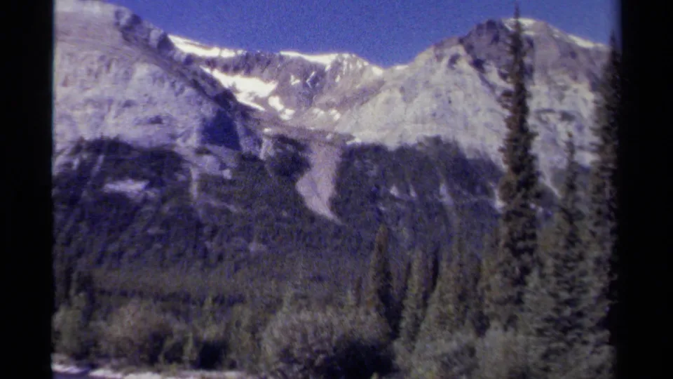 1979 British Columbia home-movie frame showing a rocky mountain ridge above dense evergreens near a river valley.