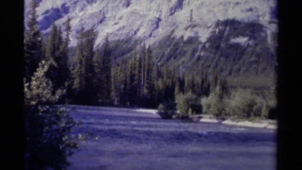 1979 8mm British Columbia landscape with a wide river, dark conifers, and pale mountain slopes behind them.