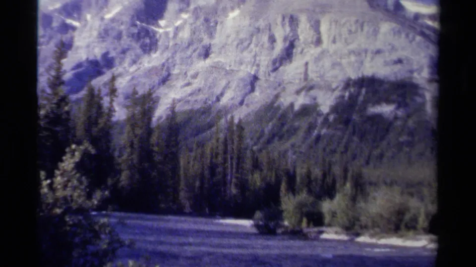 1979 British Columbia 8mm frame of a river foreground, evergreen shoreline, and steep mountain backdrop.