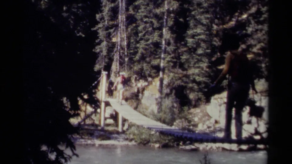 1979 British Columbia 8mm frame showing a backpacker near a narrow wooden bridge over water, surrounded by evergreen forest.