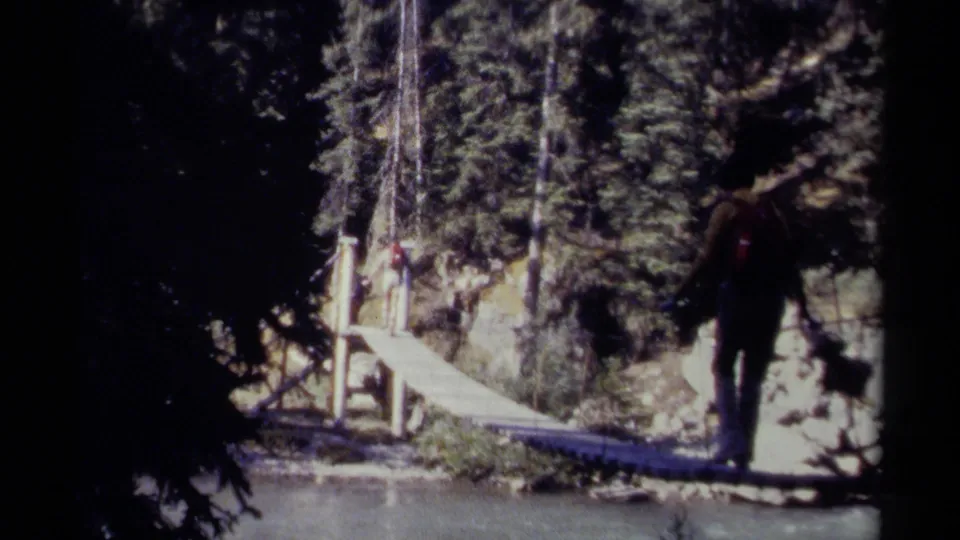 1979 British Columbia home-movie frame of a backpacker moving by a narrow wooden bridge in a forested river landscape.