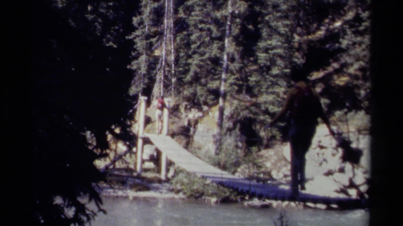 1979 British Columbia 8mm frame showing a backpacked hiker beginning to cross a narrow rope suspension bridge over a forest river.