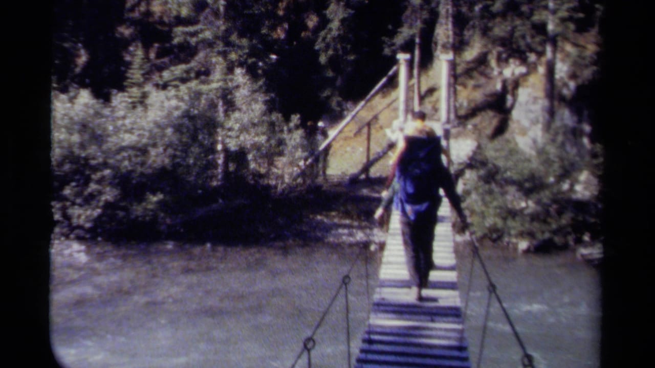 Backpacked hiker crosses a narrow suspension bridge above a river.