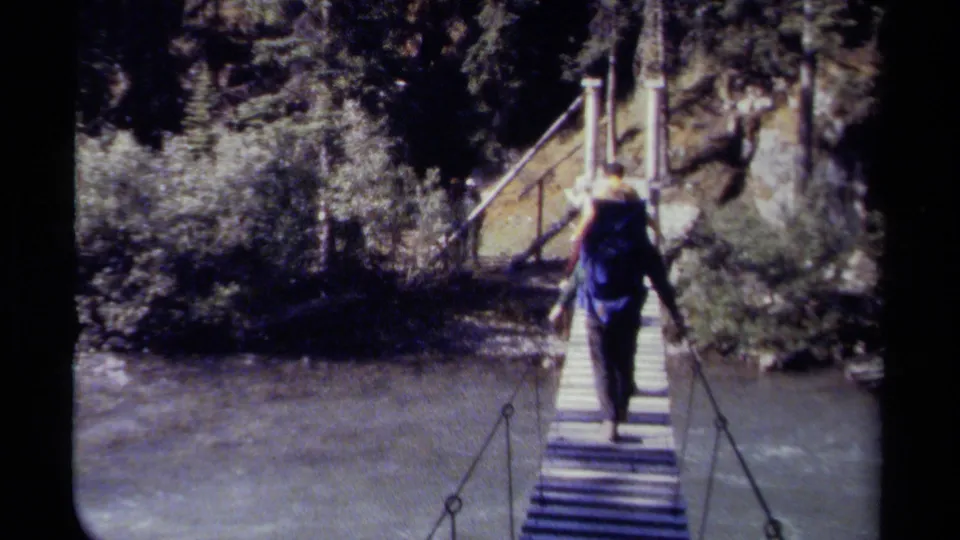 Backpacked hiker crosses a narrow rope suspension bridge over moving water in a wooded British Columbia trail setting.