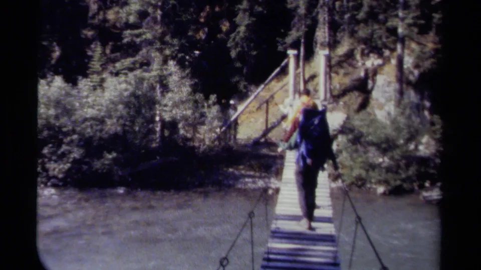 A hiker with a blue backpack continues across the suspension bridge, with dense trees and river water visible around the crossing.