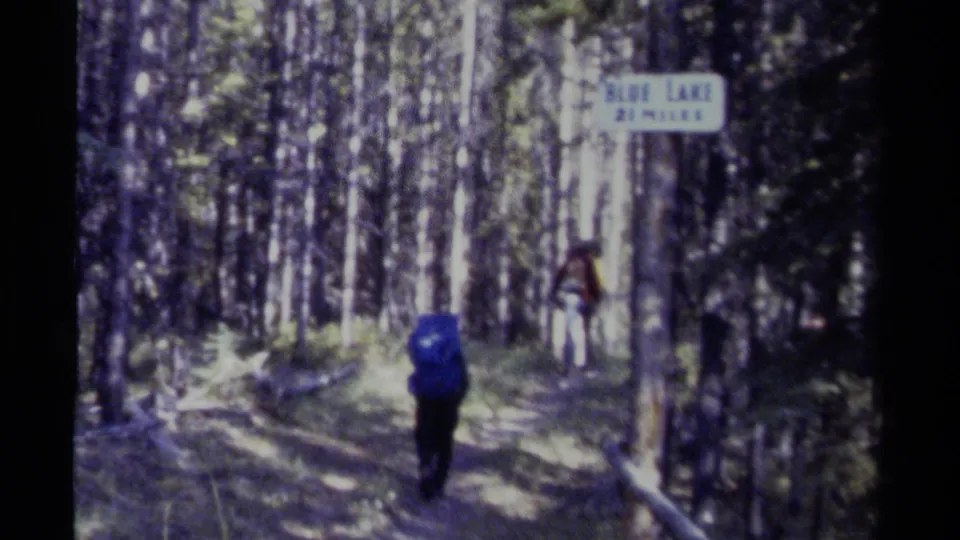 Backpacked hiker walks along a forest trail beside a sign that appears to read Blue Lake 2 Miles.