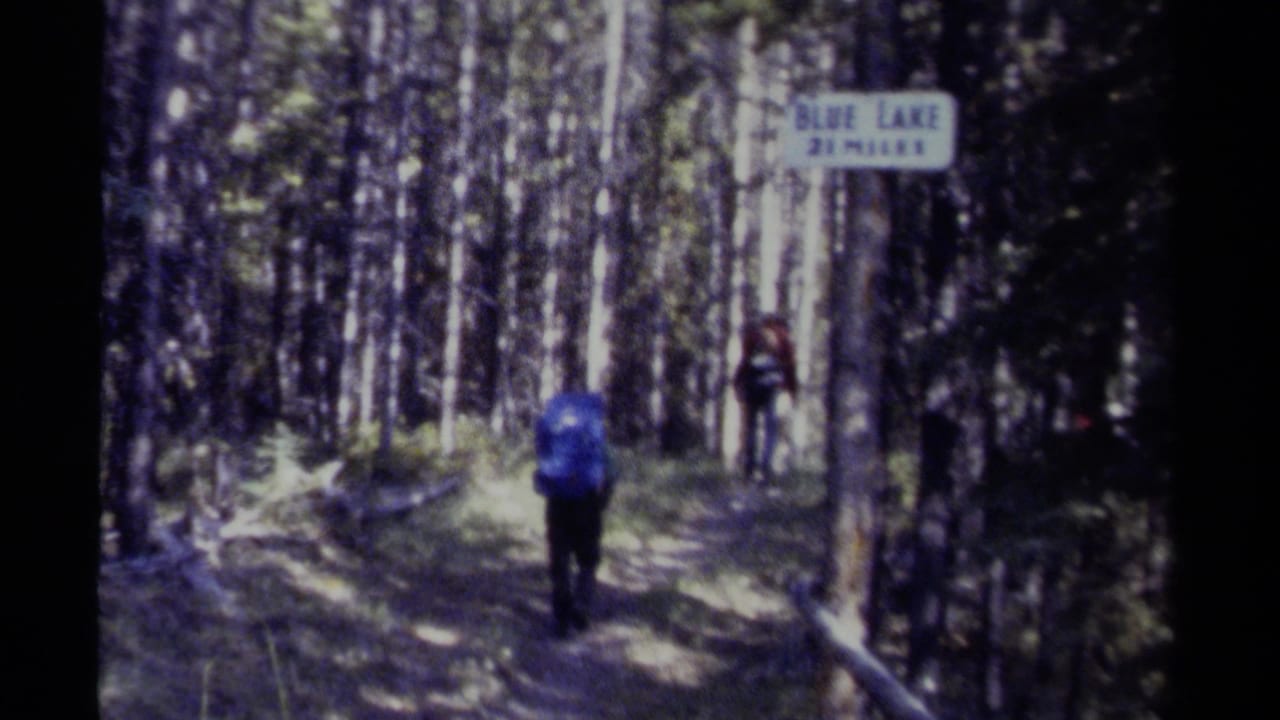 Hikers with backpacks walk away on a forest trail beside a Blue Lake two-mile sign.