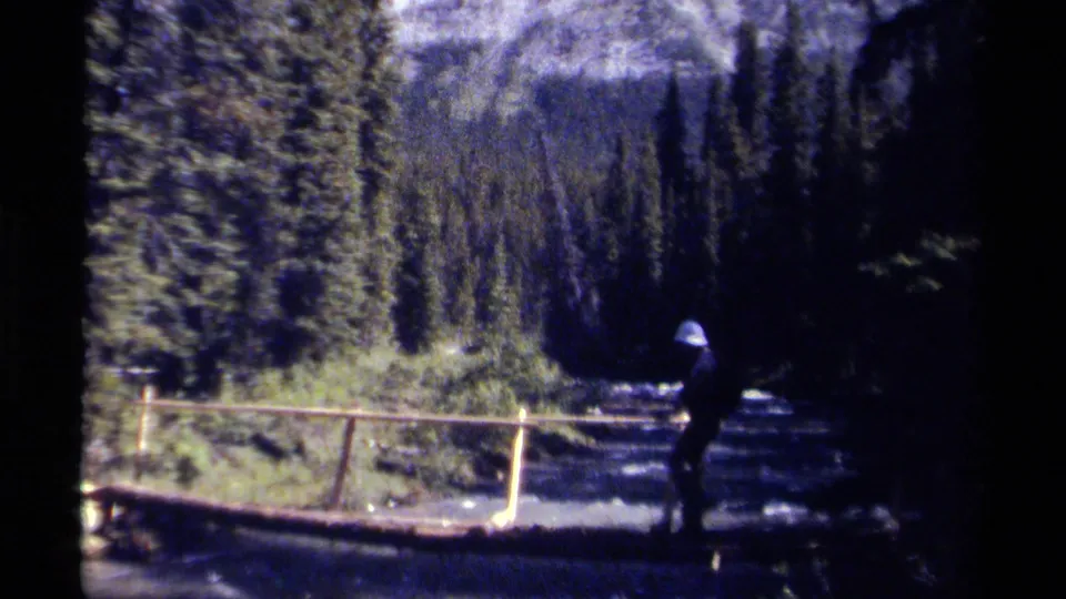 1979 British Columbia 8mm frame of a hiker crossing a wooden footbridge below evergreens and rugged mountain slopes.
