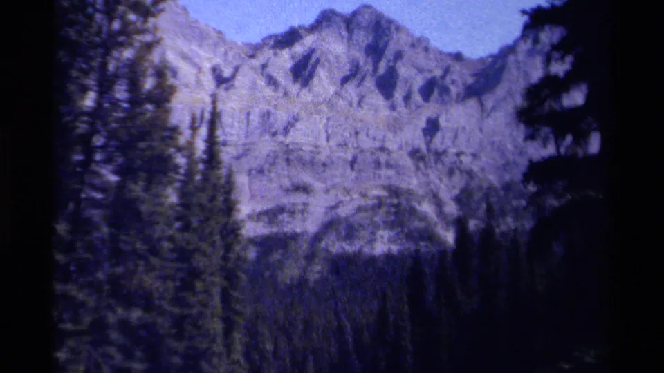 Late-1970s British Columbia home-movie frame of rugged mountain slopes and dense conifers, with the hiker nearly out of frame.