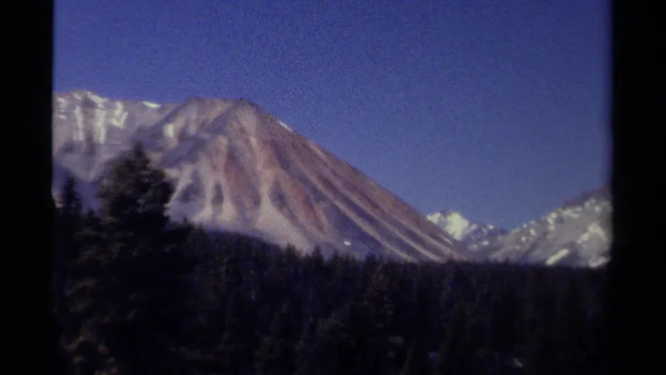 Closer 1979 8mm view of a British Columbia mountain slope, snow patches, dark forest, and clear blue sky.