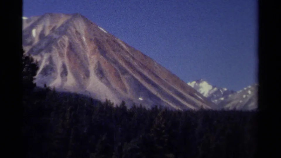 1979 British Columbia 8mm frame showing a large snow-streaked mountain slope, evergreen forest, and distant snowy peaks.