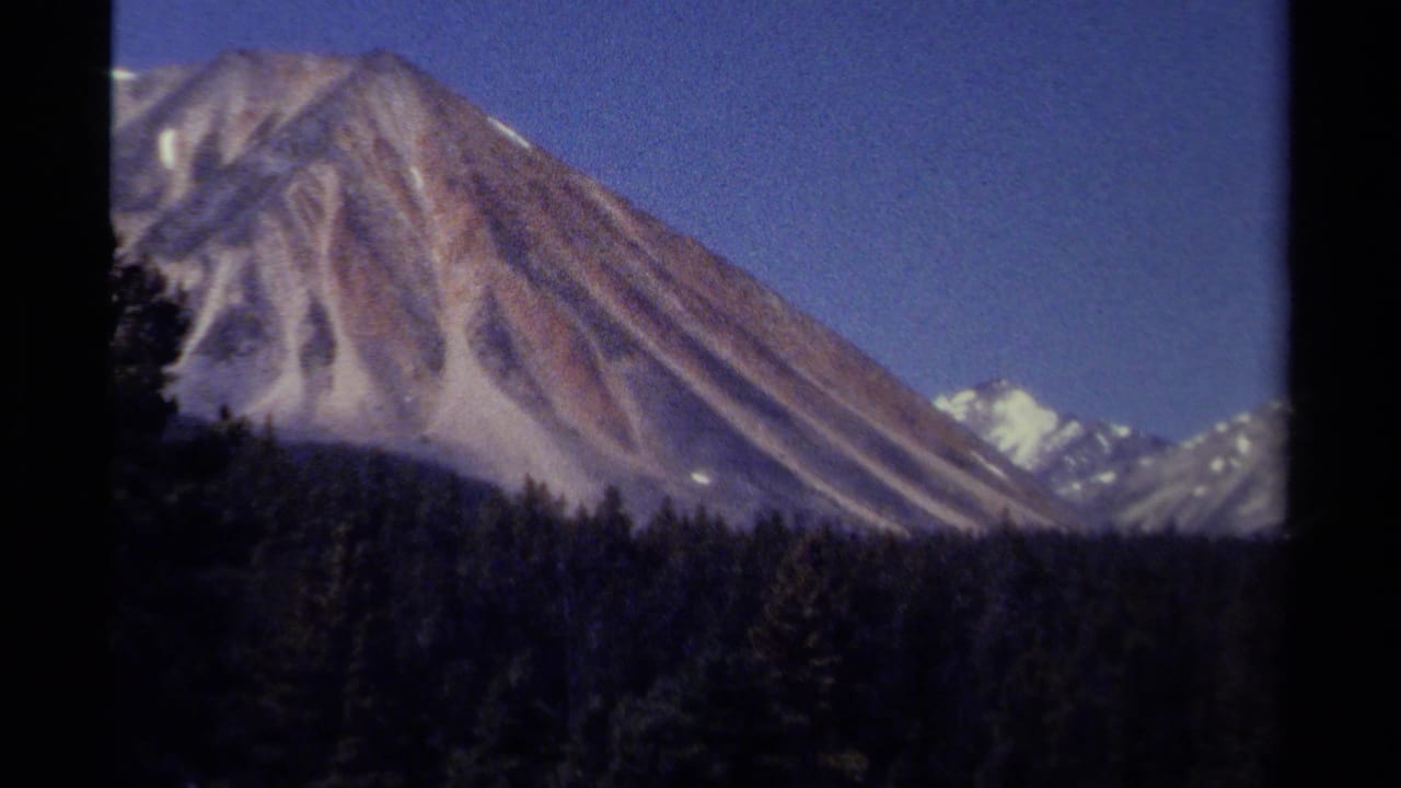 1979 8mm British Columbia mountain view with a snow-streaked peak, distant ridges, dark forest, and clear blue sky.