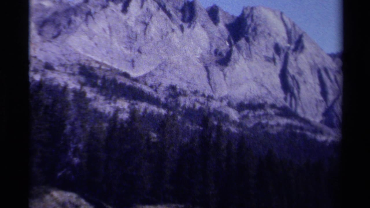 Pale rocky mountain face above a dark forested slope, framed by black film borders.