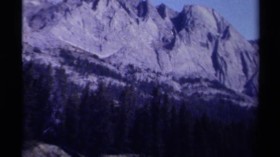 1979 8mm British Columbia mountain footage showing a pale rocky face, forested lower slope, and blue sky within the film frame.