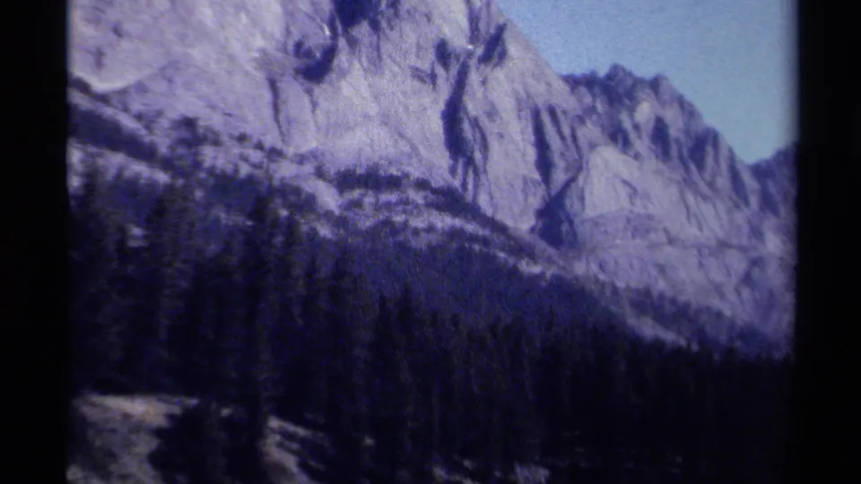 1979 British Columbia 8mm frame with a pale mountain wall, dark evergreen forest, and clear sky in a brief scenic landscape view.