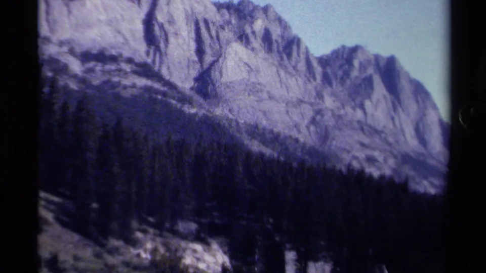 British Columbia mountain landscape on 8mm film, showing pale sky, steep rock, and a shadowed pine forest.