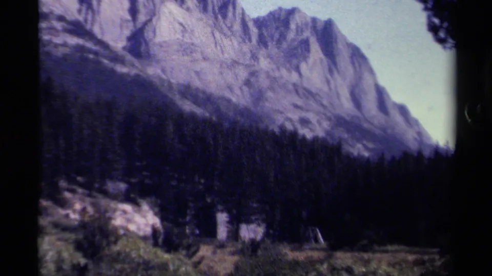 1979 home-movie frame from British Columbia showing mountain rock, pine trees, grassy ground, and a possible campsite edge.
