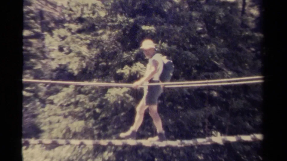 A young man in a white shirt, shorts, and light hat carefully steps along a rope bridge in a wooded British Columbia setting.