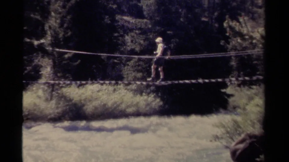 The figure continues across the suspended rope bridge over a bright, rushing river, framed by trees and shadowed riverbank.