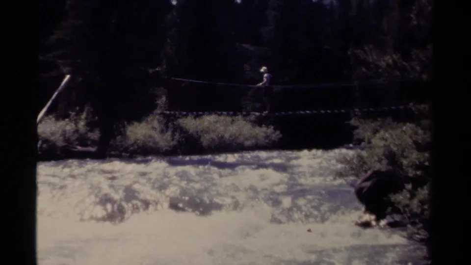 A distant figure crosses the rope bridge high over fast water, with dark forest behind and overexposed river highlights below.
