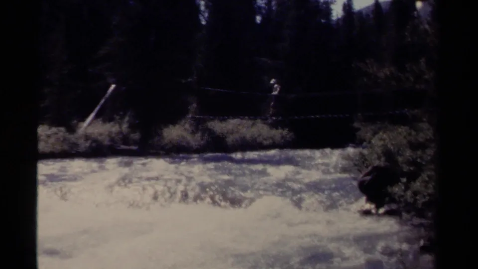 1979 British Columbia 8mm frame showing a distant person on a rope bridge over fast river water in dense forest.