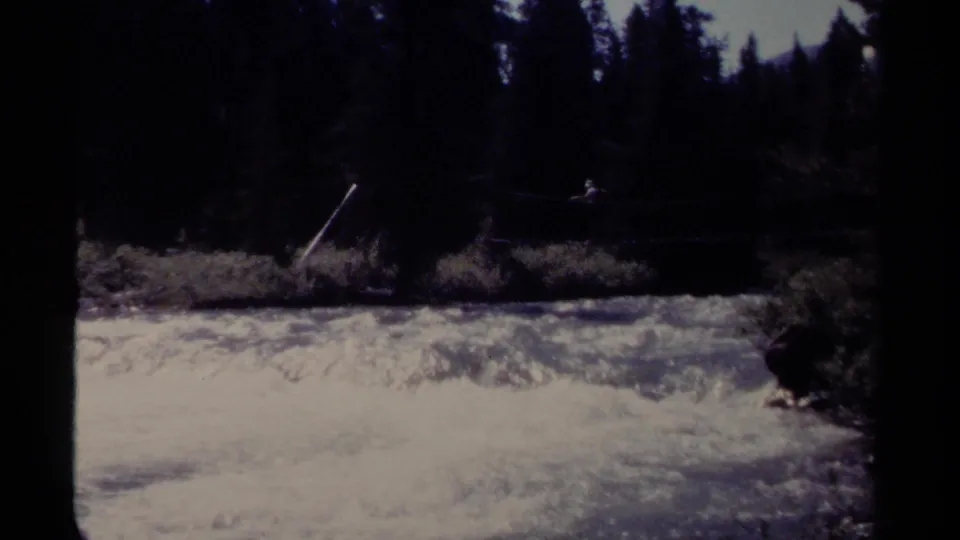 1979 British Columbia 8mm frame with a small figure on a rope bridge spanning a fast river below forested banks.
