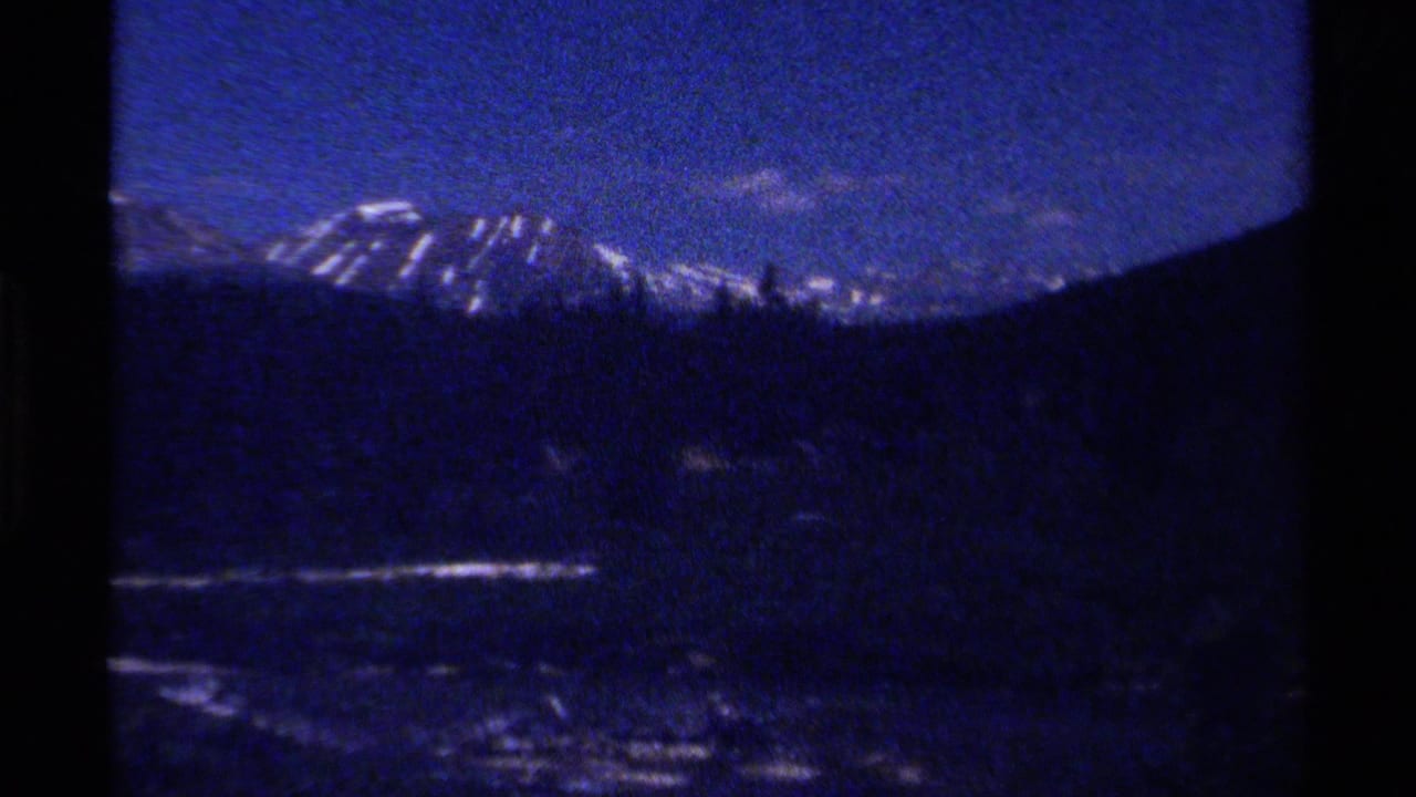 1979 British Columbia 8mm frame showing a dim forested valley, winding stream, and distant snow-streaked mountains under blue sky.
