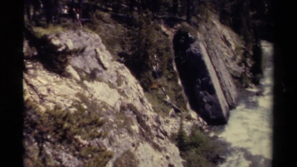 Wider British Columbia valley view on 8mm, with steep rock, evergreen trees, pale water, and a dark vertical object near the stream.
