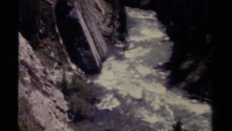 Elevated 8mm view of rushing river water passing a large dark rock in a British Columbia canyon.