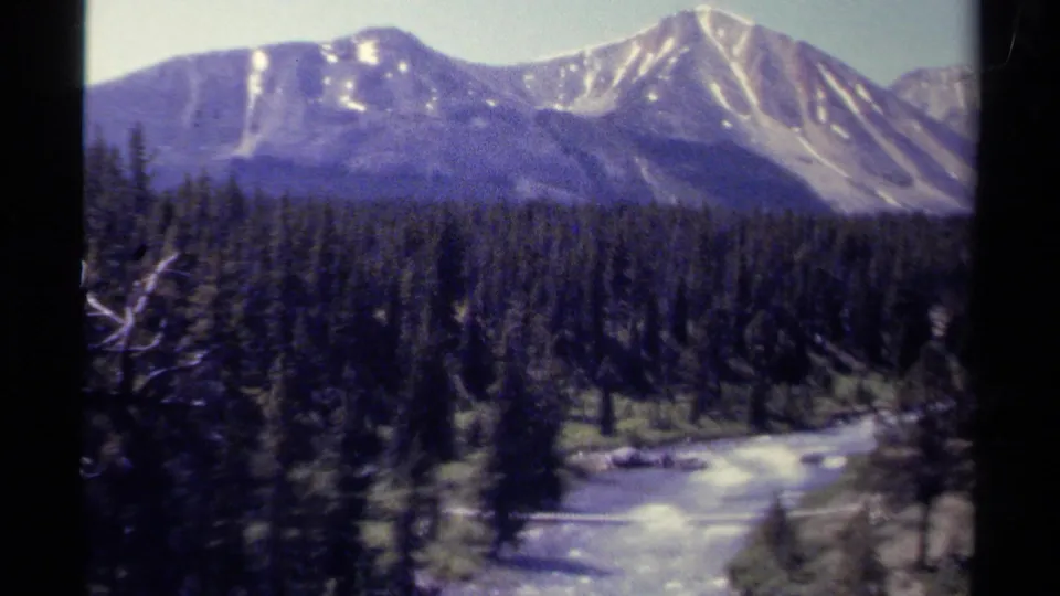 1979 8mm view of British Columbia mountain peaks, dark evergreen forest, and a river curving through the valley.