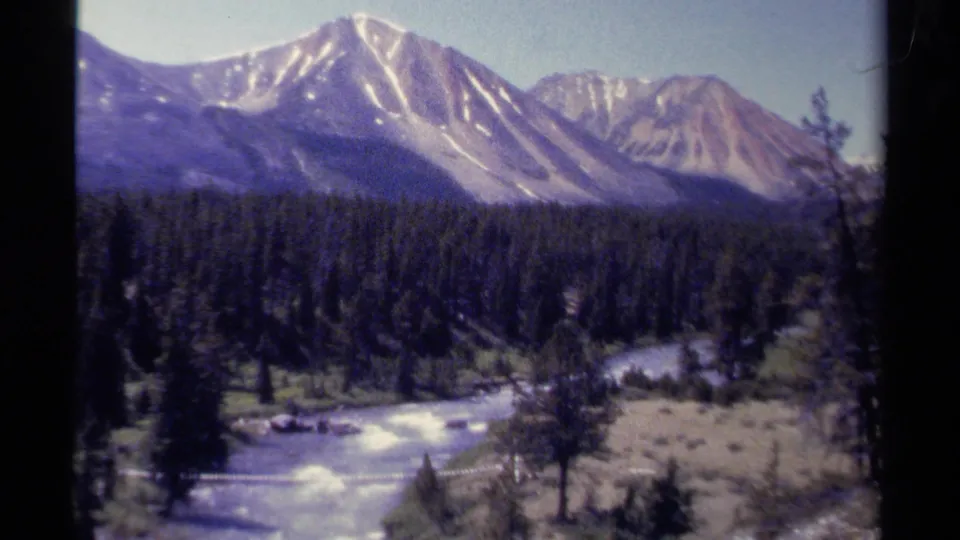 8mm home-movie frame of British Columbia mountains beyond a dark forest, with a river and light-toned banks in front.