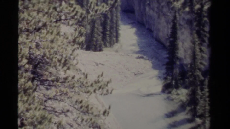 Archival 8mm frame from British Columbia showing a snow-covered river bend between conifers and shaded canyon walls.