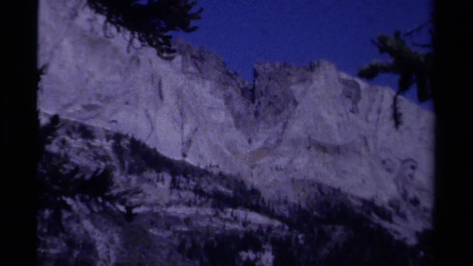 1979 8mm British Columbia landscape frame with a pale mountain ridge rising above a dark forested slope under a blue sky.