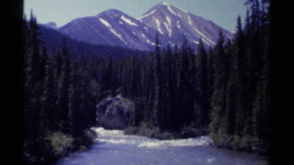 1979 8mm British Columbia frame showing snow-streaked mountain peaks beyond dense conifers with bright water or marshy ground in the foreground.