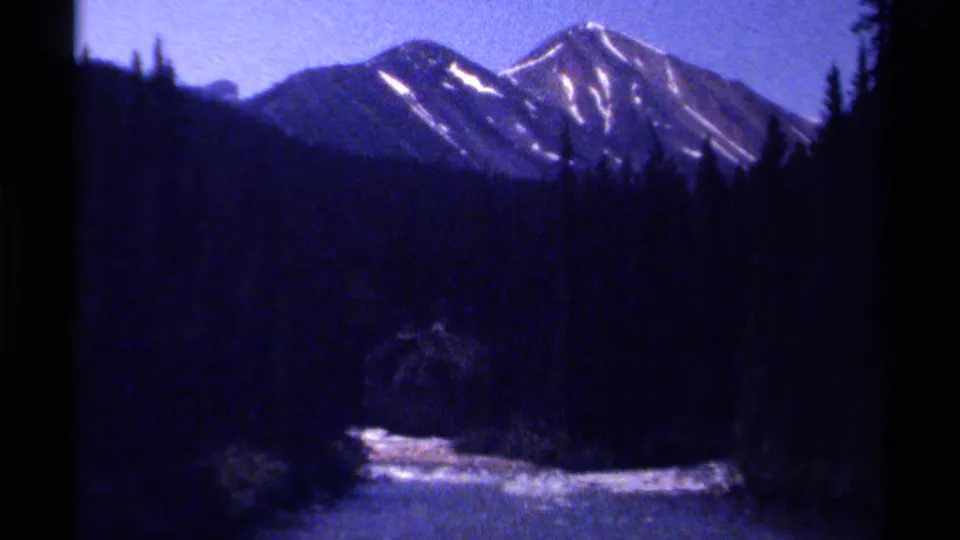1979 8mm British Columbia home-movie frame with distant snow-streaked peaks above a dark evergreen valley and a bright water channel near the bottom edge.