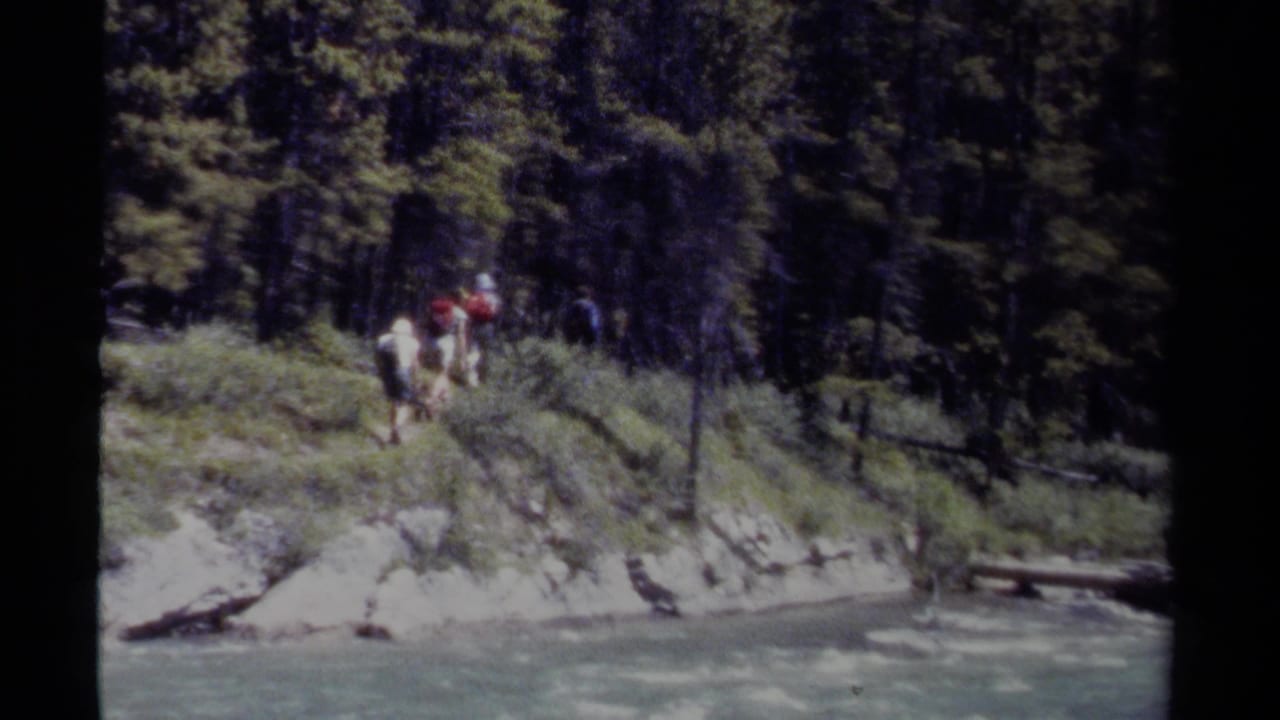 1979 British Columbia 8mm frame showing hikers with packs moving along a steep grassy bank beside a rushing river.